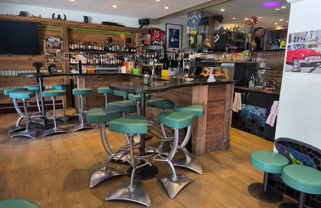 Blick auf die Overtime Bar Theke mit Barhocker und Backbar. Im Hintergrund sieht man ins Fumoir.